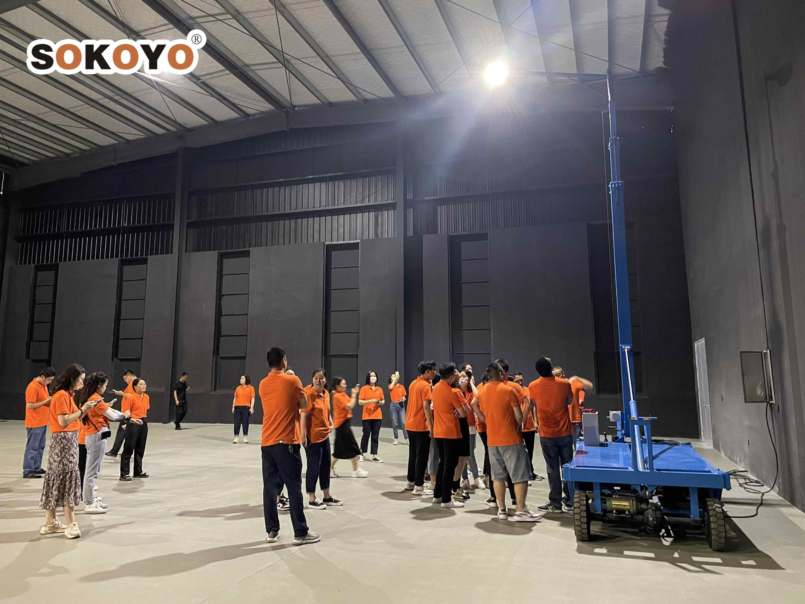 Factory workers in orange shirts gather around a blue piece of equipment in a spacious industrial facility illuminated by overhead lights