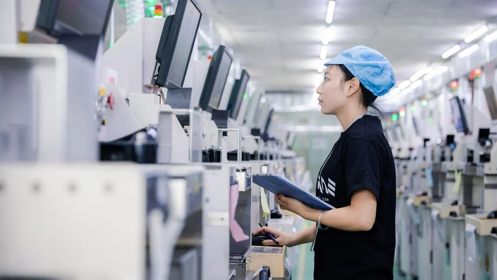 A woman wearing a blue hat stands in front of a computer focused on her work in a professional setting